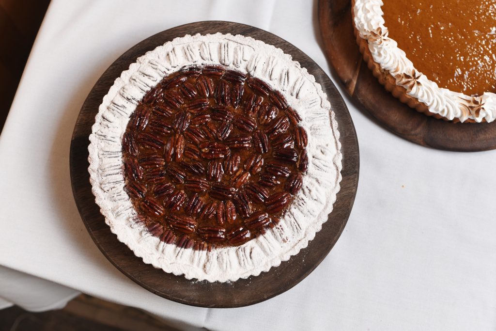 Overhead shot of a beautifully baked pecan pie on a wooden platter. The pie is topped with glossy, caramelized pecans, arranged in a symmetrical pattern, and bordered with a dusting of powdered sugar. In the background, a second pie with a golden, smooth filling and piped cream can be seen, offering a perfect contrast of textures.