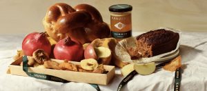 A spread of Panzer's rosh hashanah products, pomegranate, challah bread, honey, honey cake and apples all on a white tablecloth with soft lighting