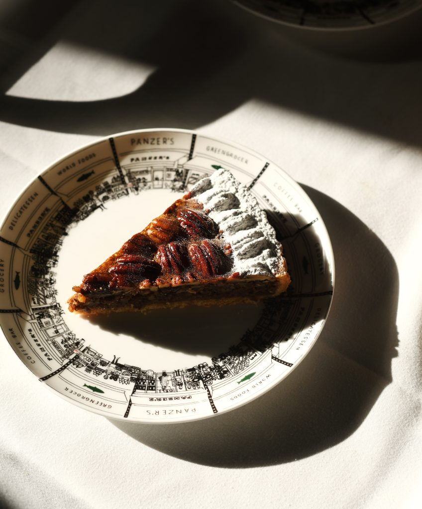 A single slice of pecan pie on a vintage-style Panzer's plate, placed on a white tablecloth. The pie features a glossy layer of caramelized pecans and a dusting of powdered sugar on the crust edge. Sunlight casts dramatic shadows, highlighting the rich texture of the pecans and the intricate illustration of the plate's design, which showcases Panzer's deli and surrounding shops.