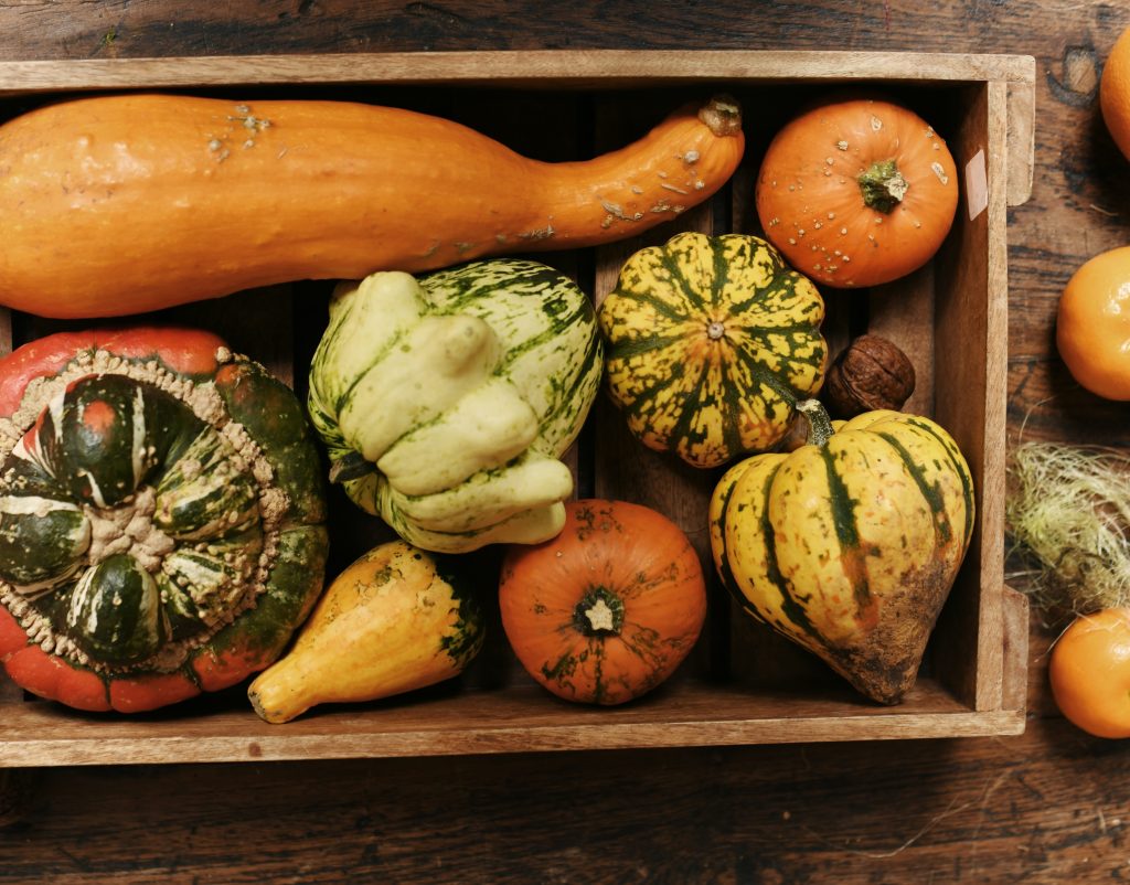 A rustic wooden crate filled with a variety of colorful pumpkins and gourds. The assortment includes orange, green, and yellow squash in different shapes and textures, showcasing the natural autumn harvest. The crate is placed on a wooden surface, with a few more pumpkins and natural elements scattered around