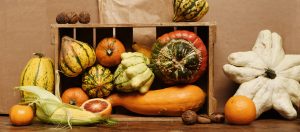 A rustic wooden crate filled with a variety of colorful pumpkins and gourds. The assortment includes orange, green, and yellow squash in different shapes and textures, showcasing the natural autumn harvest. The crate is placed on a wooden surface, with a few more pumpkins and natural elements scattered around