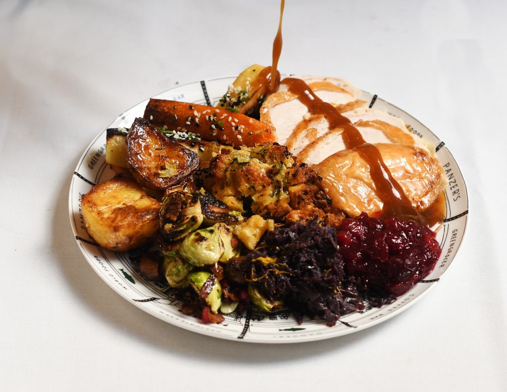 A generous Thanksgiving plate featuring sliced roast turkey breast drizzled with rich gravy, crispy roast potatoes, glazed carrots and parsnips, Brussels sprouts, homemade stuffing, braised red cabbage, and a side of cranberry sauce. The hearty meal is served on a Panzer's plate, with each element beautifully arranged for a festive feast.