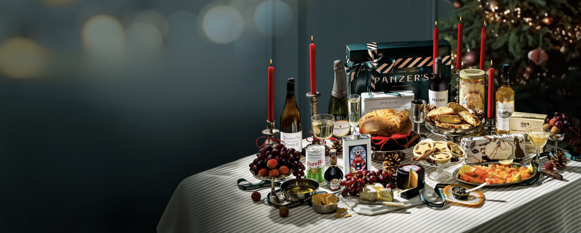 A festive spread of Panzer’s 2024 Christmas hampers, arranged on a striped tablecloth with an array of gourmet delights perfect for holiday celebrations. The setup includes items such as fine wines, cheeses, fresh fruit, and specialty canned goods, elegantly presented alongside red candles and vintage glassware. Notable products include a selection of premium biscuits, smoked salmon, and Christmas treats like panettone and mince pies, all beautifully packaged and wrapped with Panzer’s ribbon—ready for gifting or enjoying with friends and family over the holiday season.