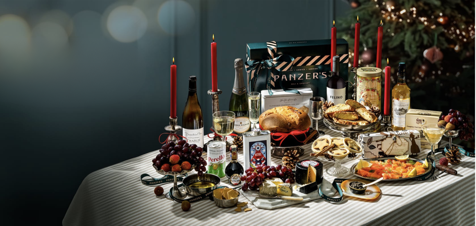 A festive spread of Panzer’s 2024 Christmas hampers, arranged on a striped tablecloth with an array of gourmet delights perfect for holiday celebrations. The setup includes items such as fine wines, cheeses, fresh fruit, and specialty canned goods, elegantly presented alongside red candles and vintage glassware. Notable products include a selection of premium biscuits, smoked salmon, and Christmas treats like panettone and mince pies, all beautifully packaged and wrapped with Panzer’s ribbon—ready for gifting or enjoying with friends and family over the holiday season.