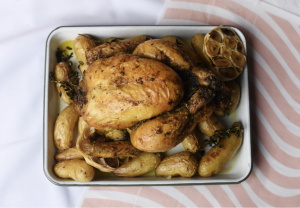 Golden rotisserie chicken placed on a white tray, surrounded by roasted fingerling potatoes, garlic bulbs, and sprigs of fresh herbs, with a striped pink and white background