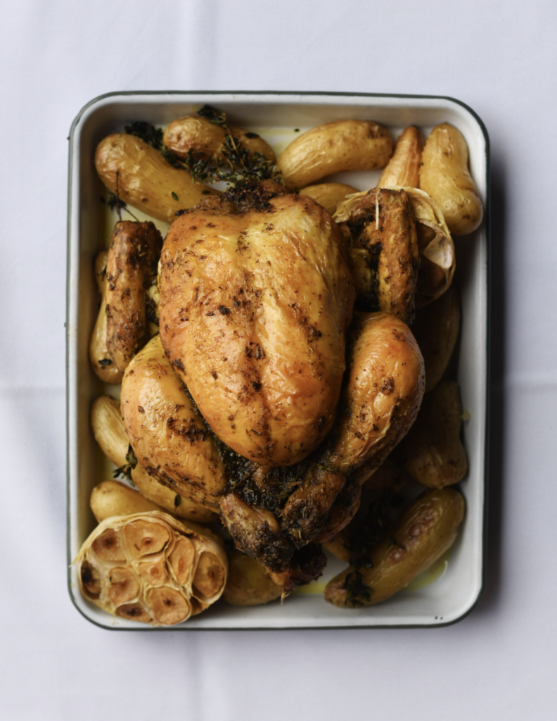 A perfectly roasted rotisserie chicken, golden and glistening, surrounded by roasted garlic bulbs and crispy potatoes in a white enamel tray, set against a simple white background.