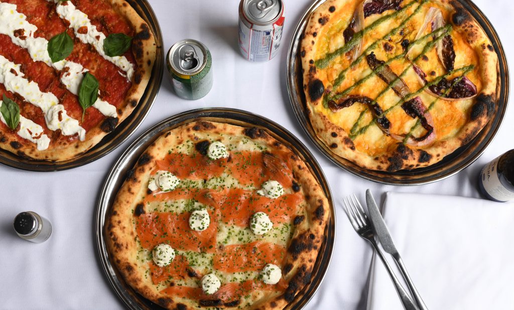 Three cooked pizzas on metal trays sit atop a white tablecloth. The first pizza has a classic tomato base with fresh basil, creamy white stracciatella, and sundried tomatoes. The second features Panzer's smoked salmon, dollops of herbed cream cheese, and finely chopped chives. The third has a golden, squash base topped with grilled radicchio, green sauce drizzles, and charred asparagus. Around the table, there are cans of soda, a bottle of beer, and neatly placed cutlery.