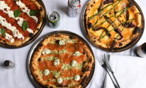 Three cooked pizzas on metal trays sit atop a white tablecloth. The first pizza has a classic tomato base with fresh basil, creamy white stracciatella, and sundried tomatoes. The second features Panzer's smoked salmon, dollops of herbed cream cheese, and finely chopped chives. The third has a golden, squash base topped with grilled radicchio, green sauce drizzles, and charred asparagus. Around the table, there are cans of soda, a bottle of beer, and neatly placed cutlery.