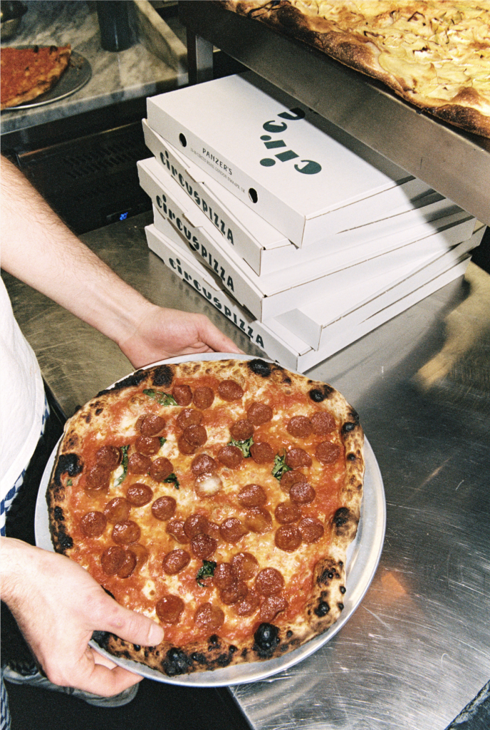 A person holds a freshly baked pepperoni pizza on a metal tray, its crust blistered and charred in spots. Behind them, a stack of white "CIRCUS PIZZA" boxes bearing the Panzer’s logo sits on a stainless steel countertop. Above, another pizza, possibly a different variety, rests on a warming shelf. The scene captures the energy of a busy kitchen, where pizzas are made fresh and ready to be boxed up for takeaway.