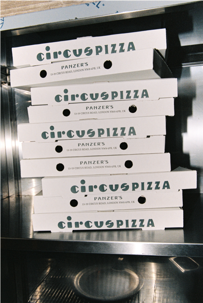 A stack of white pizza boxes labeled "CIRCUS PIZZA" sits on a stainless steel surface. Each box also features the Panzer’s logo and address: "13-19 Circus Road, London NW8 6PB, UK." The bold, stylized typography stands out against the clean design of the boxes, suggesting fresh, high-quality pizza ready for takeaway or delivery.