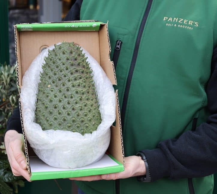 A Panzer's Deli & Grocery employee in a green uniform holds an open cardboard box containing a large soursop fruit, carefully wrapped in bubble wrap. The background features the exterior of Panzer's store with a glimpse of its warm-lit interior.