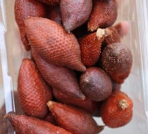 A plastic container filled with salak, also known as snake fruit. These small, reddish-brown fruits have a distinctive scaly skin that resembles snake scales. Their pointed ends and slightly rough texture give them an exotic appearance. Inside, the flesh is sweet and tangy, with a crisp yet juicy bite. A unique tropical treat!