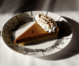 A single slice of pumpkin pie on a vintage-style Panzer's plate, placed on a white tablecloth. The pie features a glossy layer of pumpkin filling and thick cloud of blowtorched meringue on the crust edge. Sunlight casts dramatic shadows, highlighting the rich texture of the marshmallow meringue and the intricate illustration of the plate's design, which showcases Panzer's deli and surrounding shops.